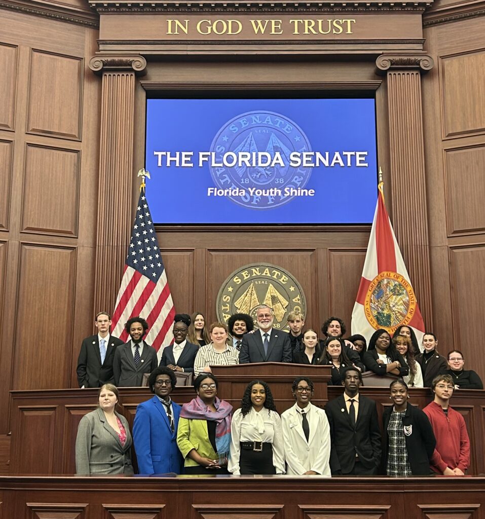 Group of diverse young people and adults in formal attire posing in front of a Florida Senate backdrop with flags and wood paneling.
